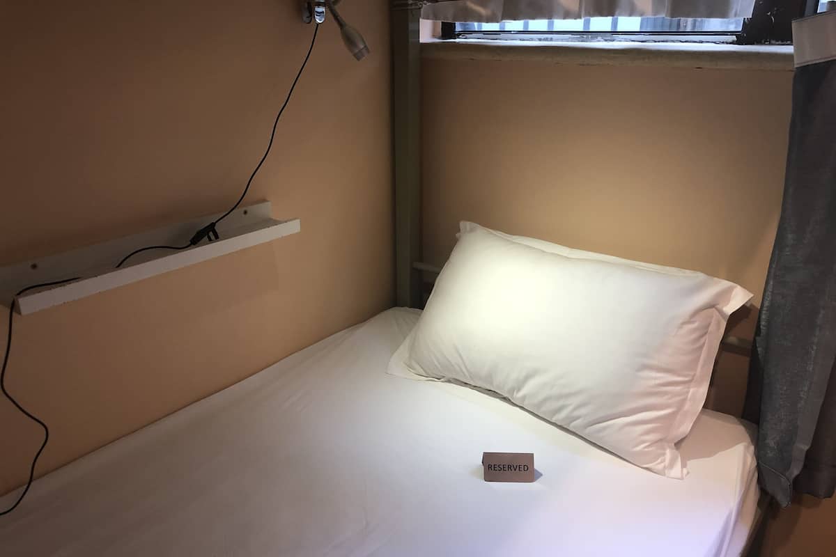A single bed with a white pillow and a 'RESERVED' sign, a power outlet, and a window above the bed.