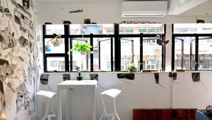 A room with a white table and two white chairs, a wooden bench, and a window with black frames and hanging plants. - Hop Inn - Hostel (Kowloon)