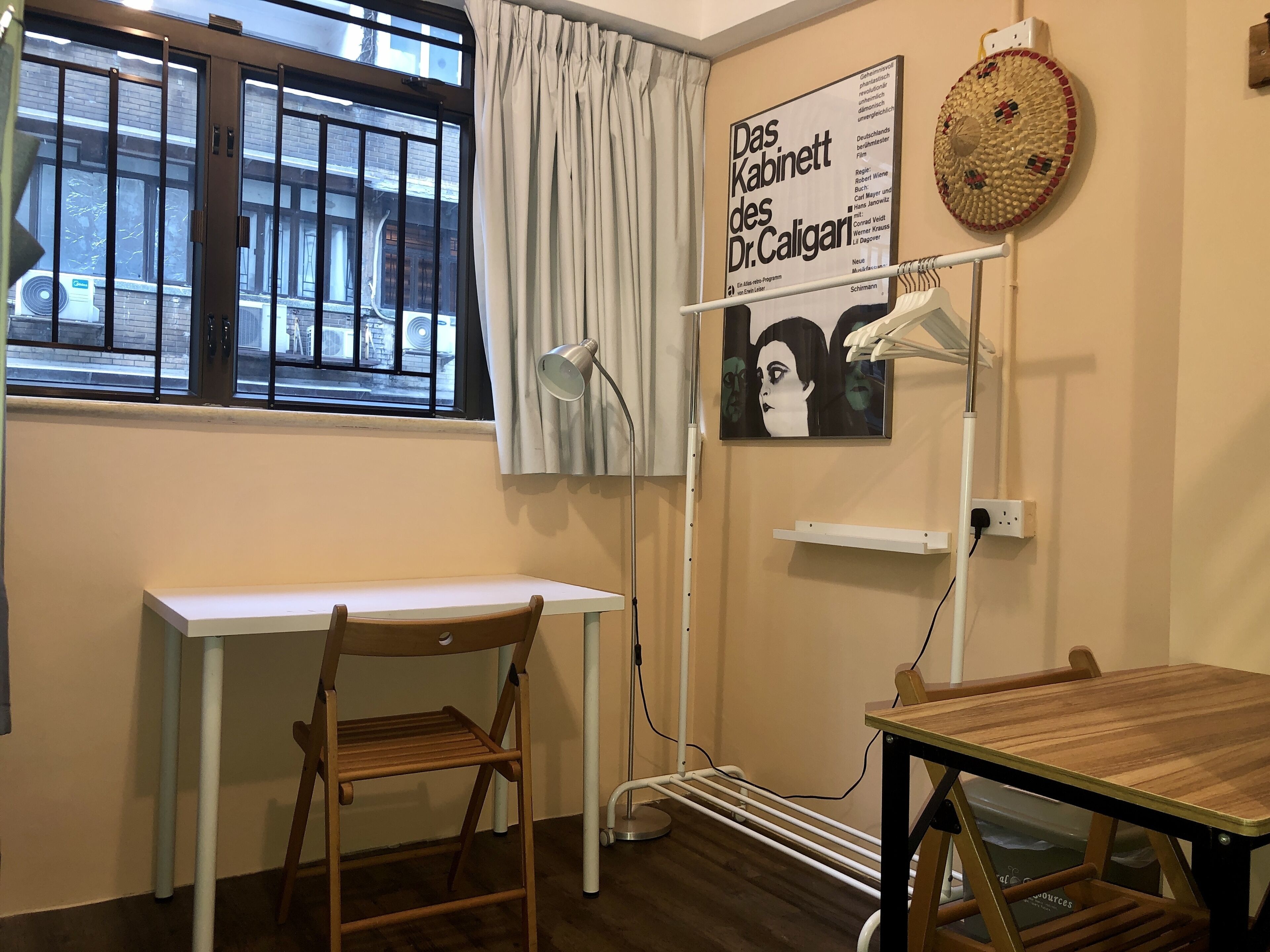 Een kamer met een wit bureau, een houten stoel, een staande lamp, een poster aan de muur en een raam met uitzicht.