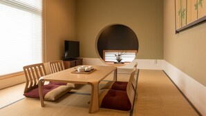 Ein traditioneller japanischer Raum mit Tatami-Boden, einem niedrigen Tisch mit Kissen und einem Fernseher auf einem Schrank.