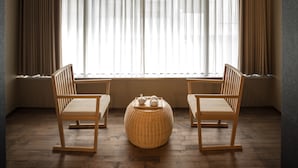 Ein Zimmer mit zwei Holzstühlen, einem gewebten Hocker und einem Fenster mit durchscheinenden Vorhängen.