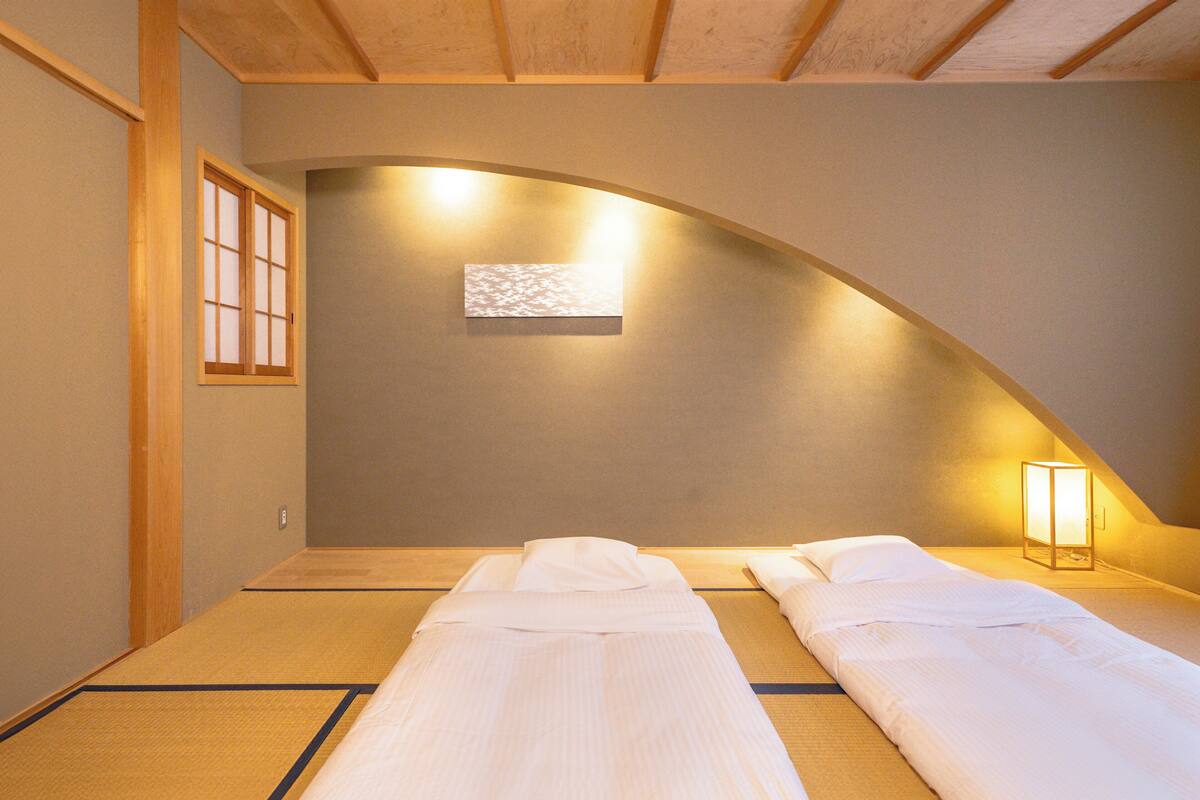 Una habitación con dos camas, suelo de tatami, techo inclinado y una lámpara fijada a la pared.