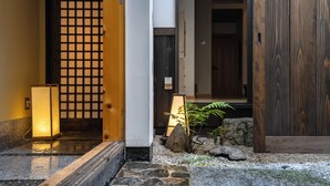 En traditionel japansk indgang med en træport, sten trapper, en lille have med sten og en plante, samt en lanterne.