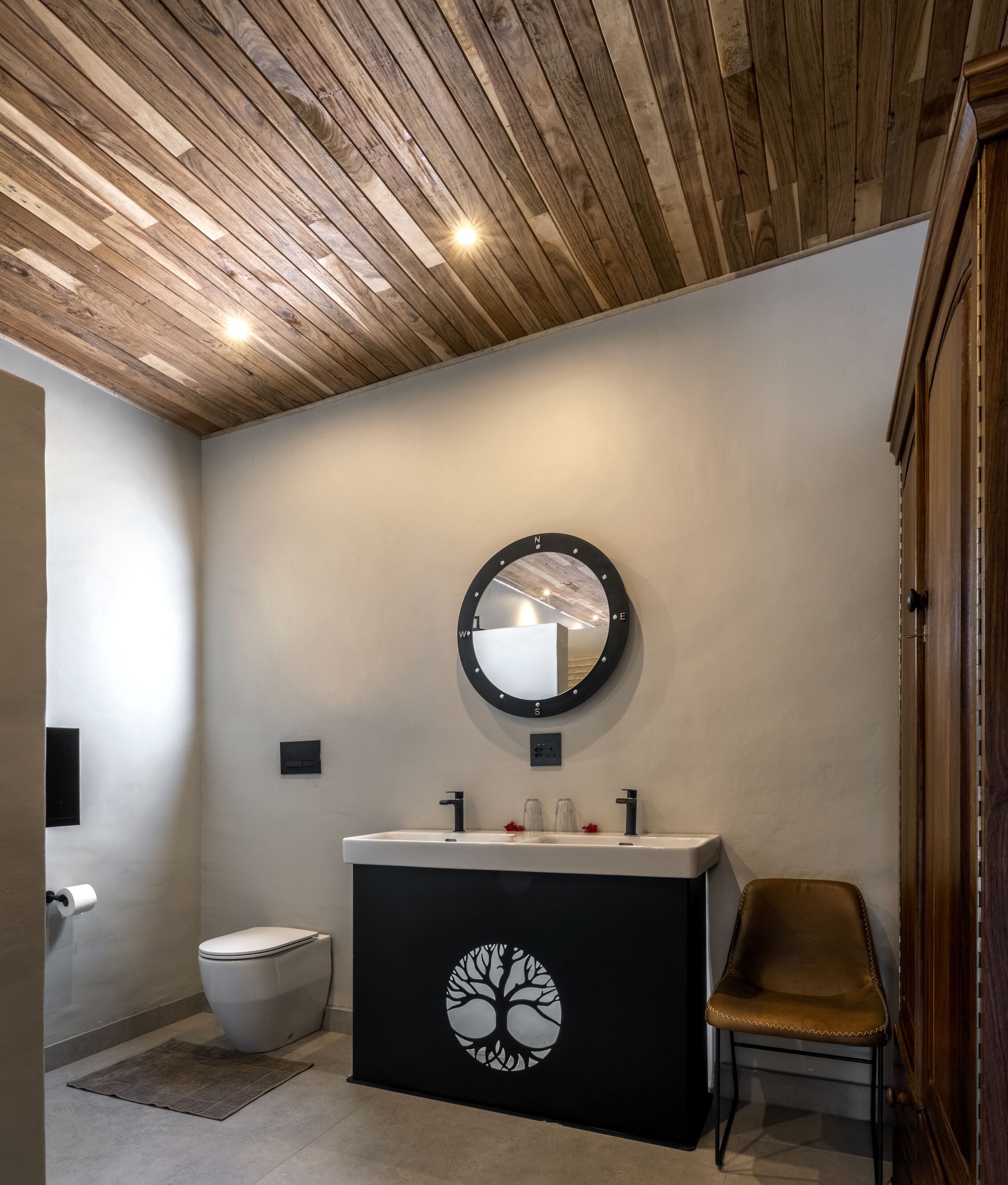 A modern bathroom with a wooden ceiling, a round mirror, a black vanity with a tree design, a white toilet, and a brown chair.