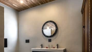 A modern bathroom with a wooden ceiling, a round mirror, a black vanity with a tree design, a white toilet, and a brown chair.