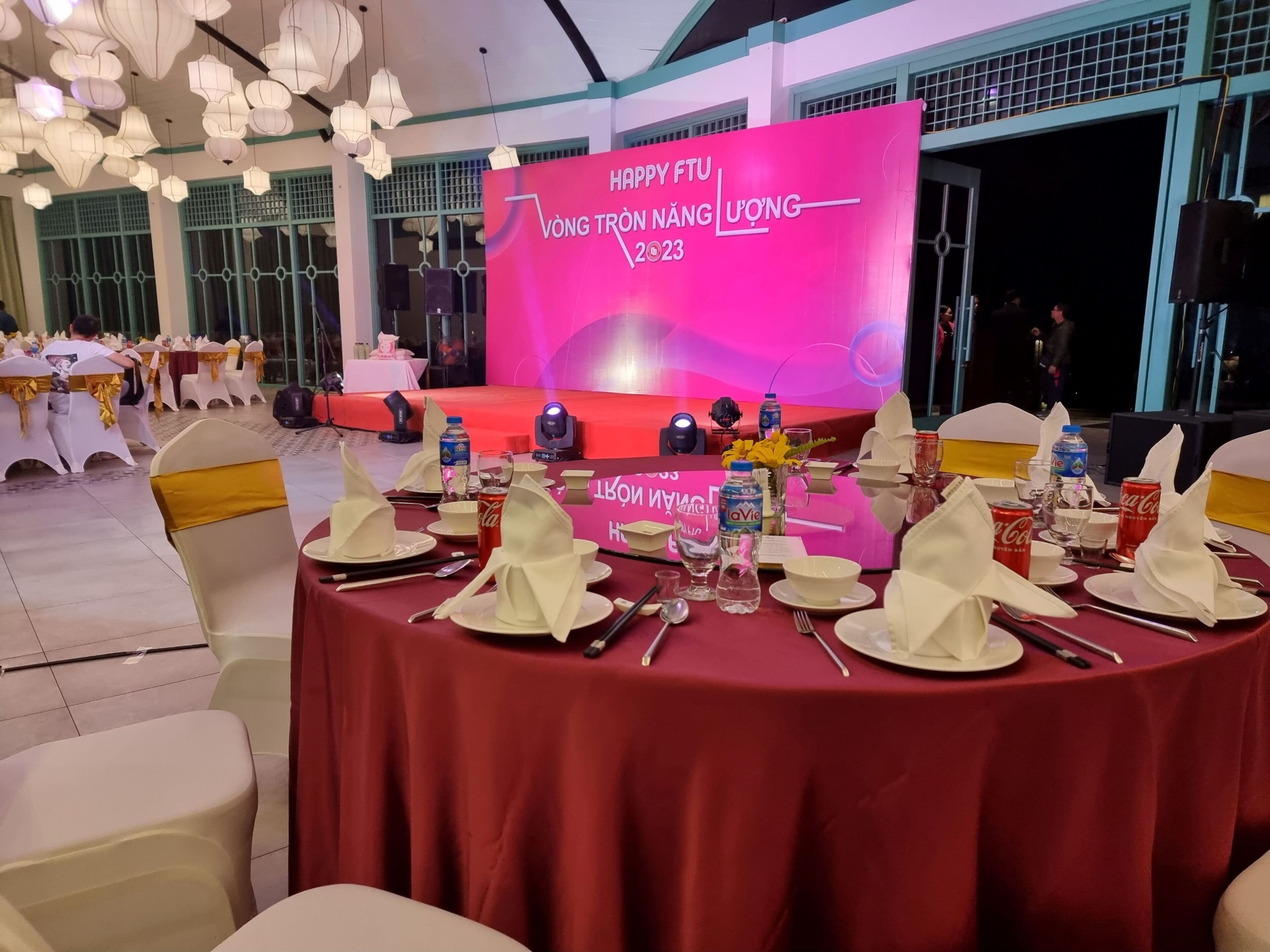 Une salle de banquet avec des tables rondes dressées pour un événement formel, comprenant une scène avec un décor de fond rose et une banderole.