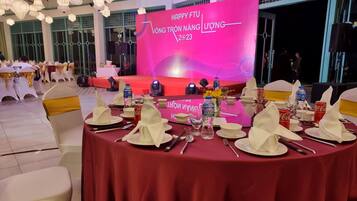 Une salle de banquet avec des tables rondes dressées pour un événement formel, comprenant une scène avec un décor de fond rose et une banderole.