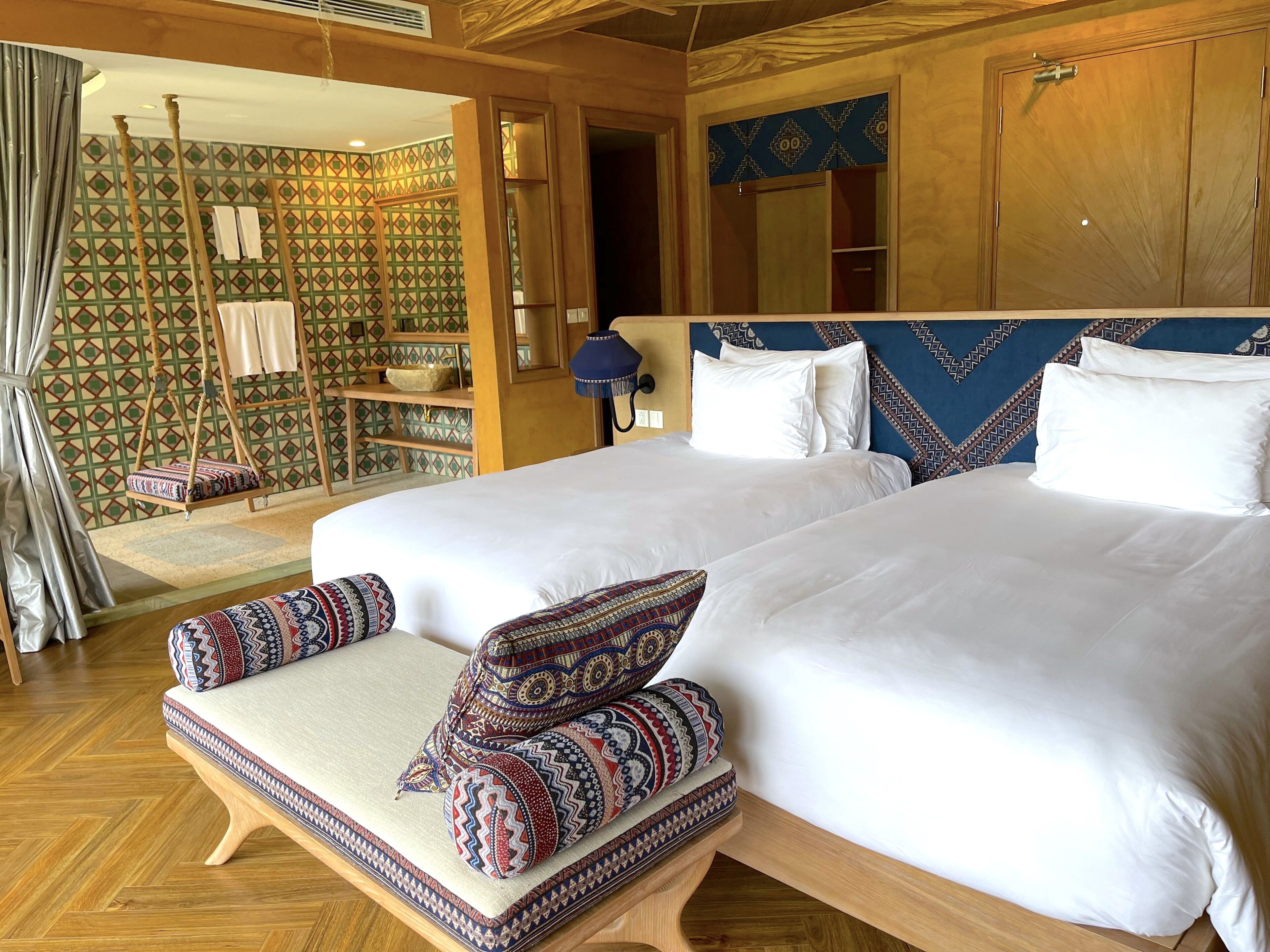 Ein Hotelzimmer mit einem großen Bett, zwei Kissen, einem Holz-Kopfschild, einem Hängesessel und einer Wand mit Muster.