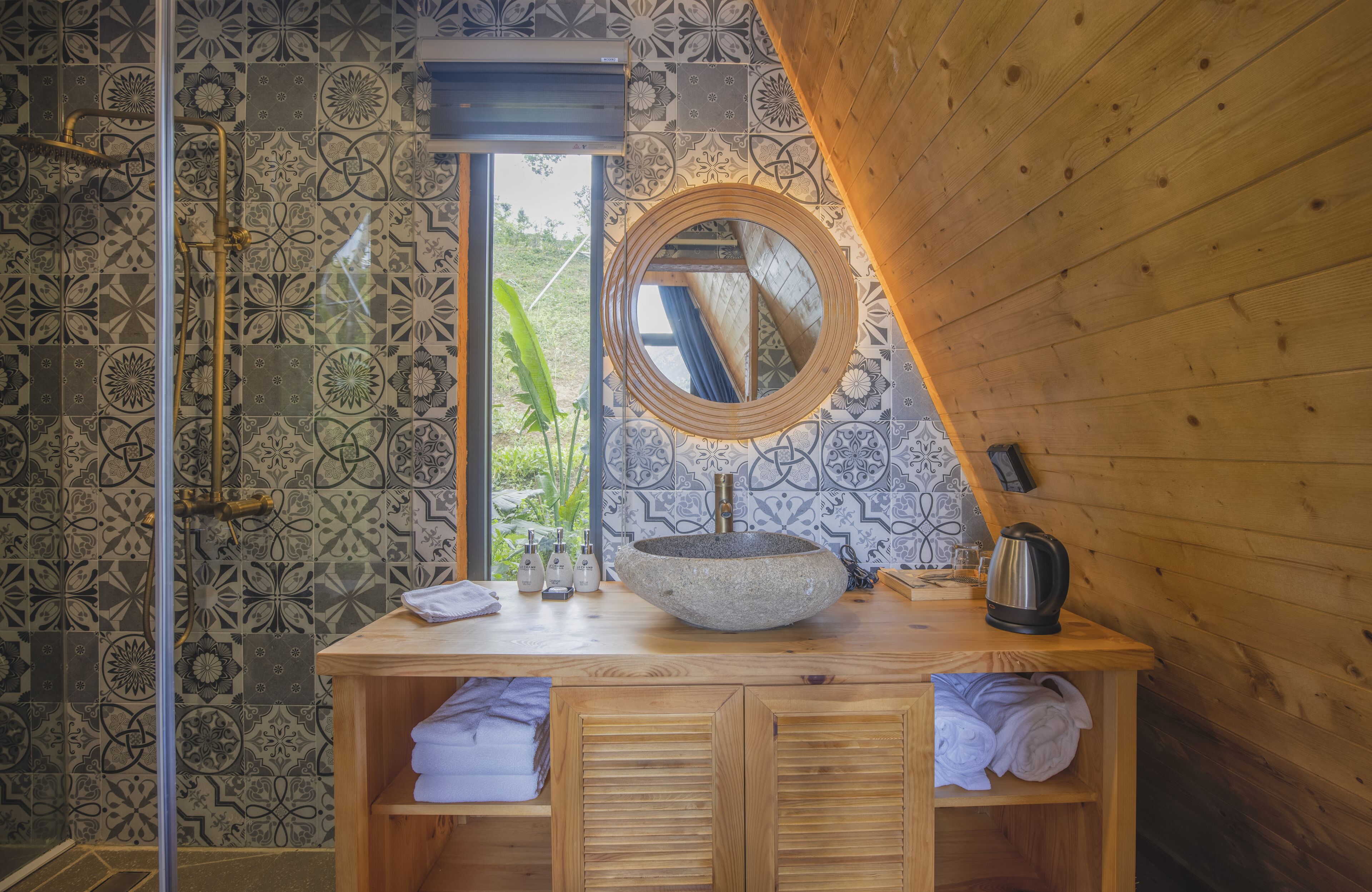 Un baño con tocador de madera, espejo redondo, lavabo de piedra y zona de ducha con azulejos estampados.