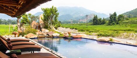 Zona de piscina con sillones reclinables, techo de paja y vistas a campos verdes y montañas.