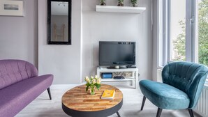 A modern living room with a purple sofa, teal armchair, and a round coffee table with a book and flowers. A flat-screen TV is on a white stand, and there's a window with a view of greenery. - Bed and Breakfast The Rose (Haarlem)