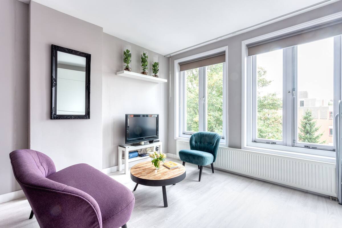 A modern living room with a large window, a flat-screen TV, a round coffee table with a vase, and two chairs.