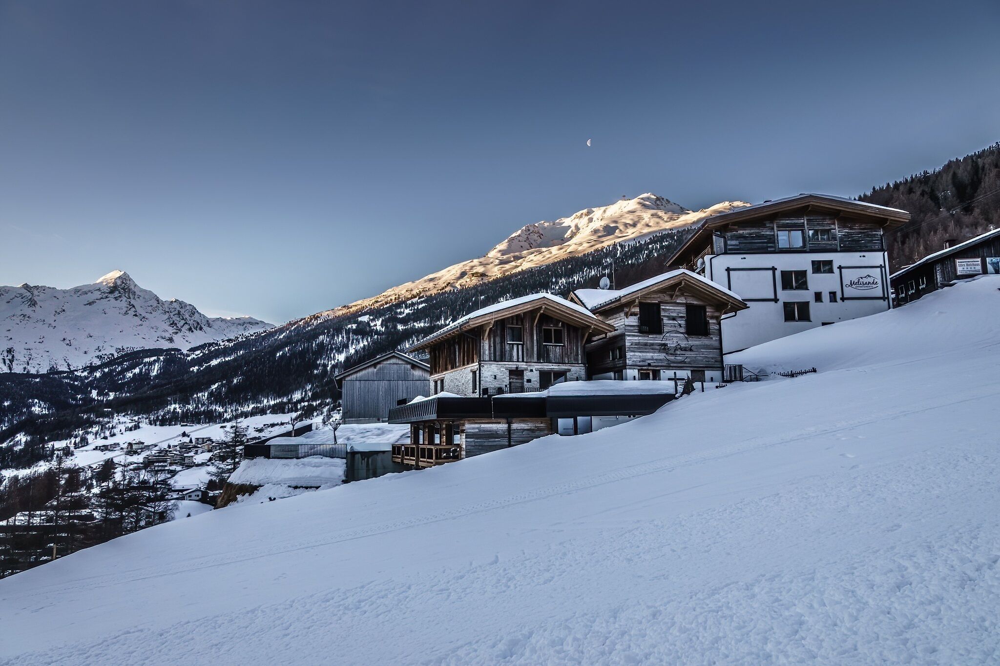 A snow-covered mountain landscape with traditional wooden buildings, including a chalet and a modern hotel with a sign reading "Naturhotel."