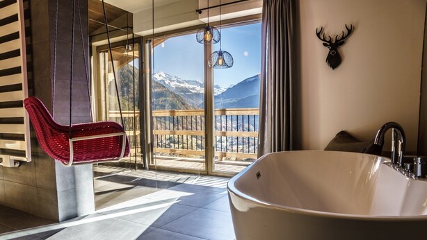 Une salle de bain moderne avec une grande baignoire, un fauteuil en osier rouge et une vue sur des montagnes enneigées, visible à travers de grandes fenêtres.