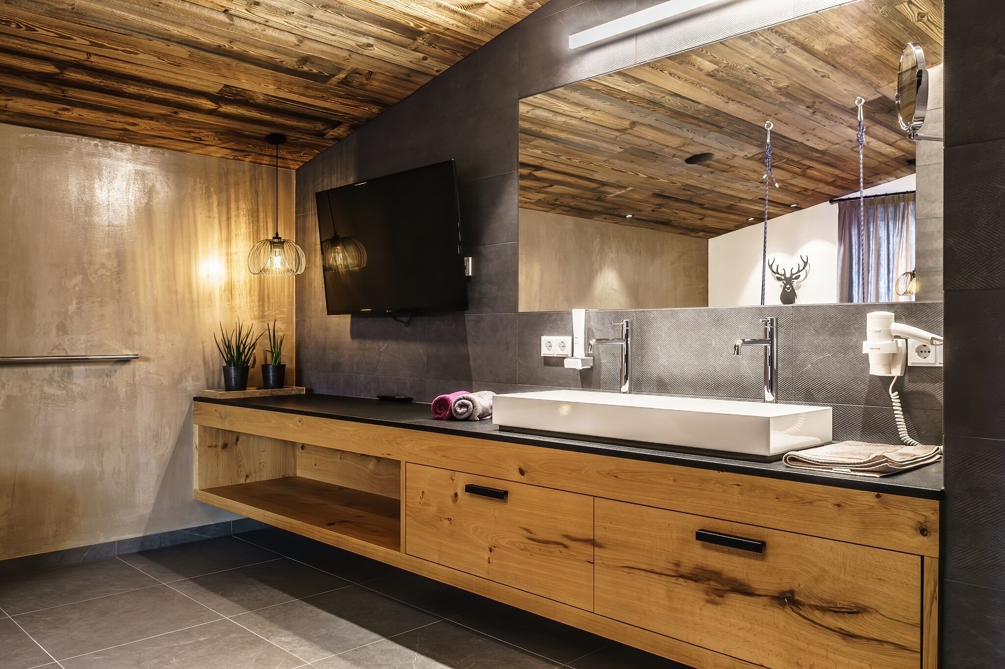 A modern bathroom with a wooden ceiling, a large mirror, a sink, and a television.