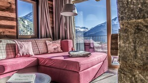 Une pièce chaleureuse avec un canapé d’angle rose, une petite table ronde sur laquelle repose un livre, et une vue sur des montagnes enneigées par de grandes fenêtres.