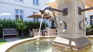 A fountain with water flowing into a pool, surrounded by outdoor furniture and a bench. - Exklusive Ferienzimmer Gut Heidefeld (Bocholt)