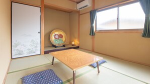 A traditional Japanese room with tatami flooring, a low wooden table, cushions, a shoji screen, and a window with curtains. - The EXP Yachiyo (Uda)
