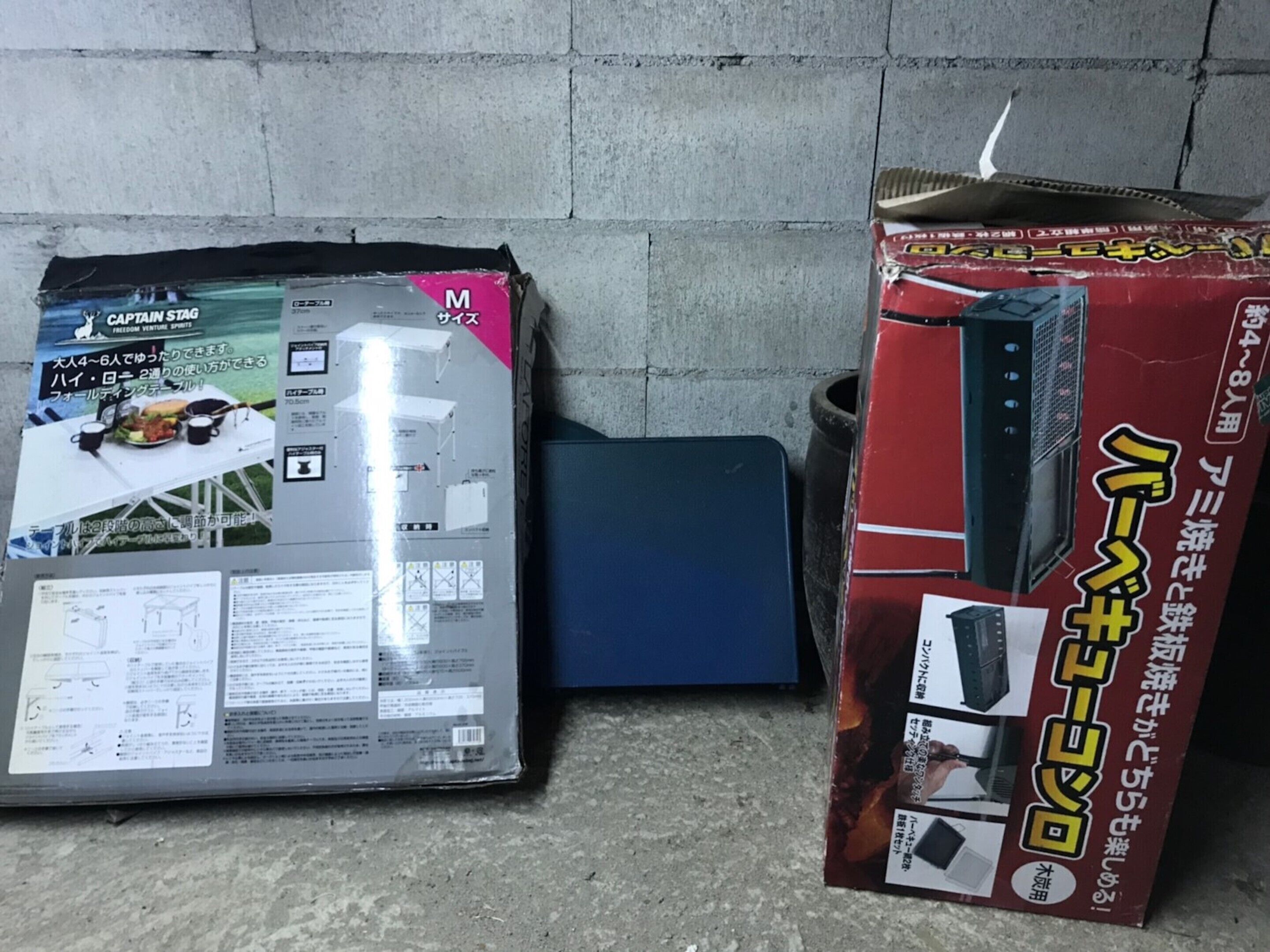 Dos cajas, una con tapa gris y otra con tapa roja, sobre un suelo de hormigón, junto a una pared.