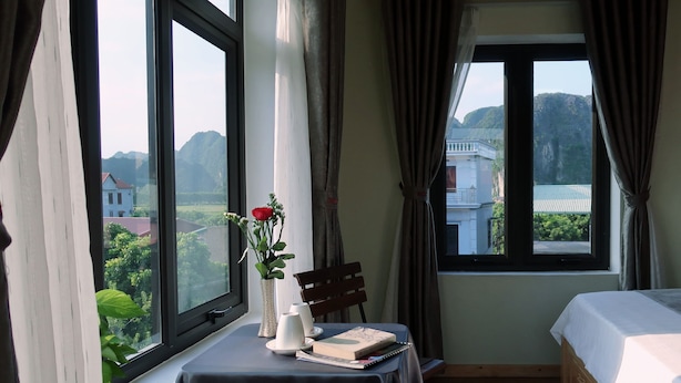 A hotel room with a bed, a table with a vase of flowers, a chair, and a view of the outside. - Tam Coc Sweet Home (Nam Hoa Lu)