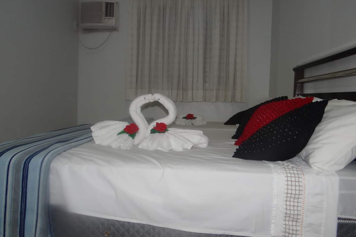 A hotel room with a bed, a window with curtains, an air conditioning unit, and a wall-mounted radiator.
