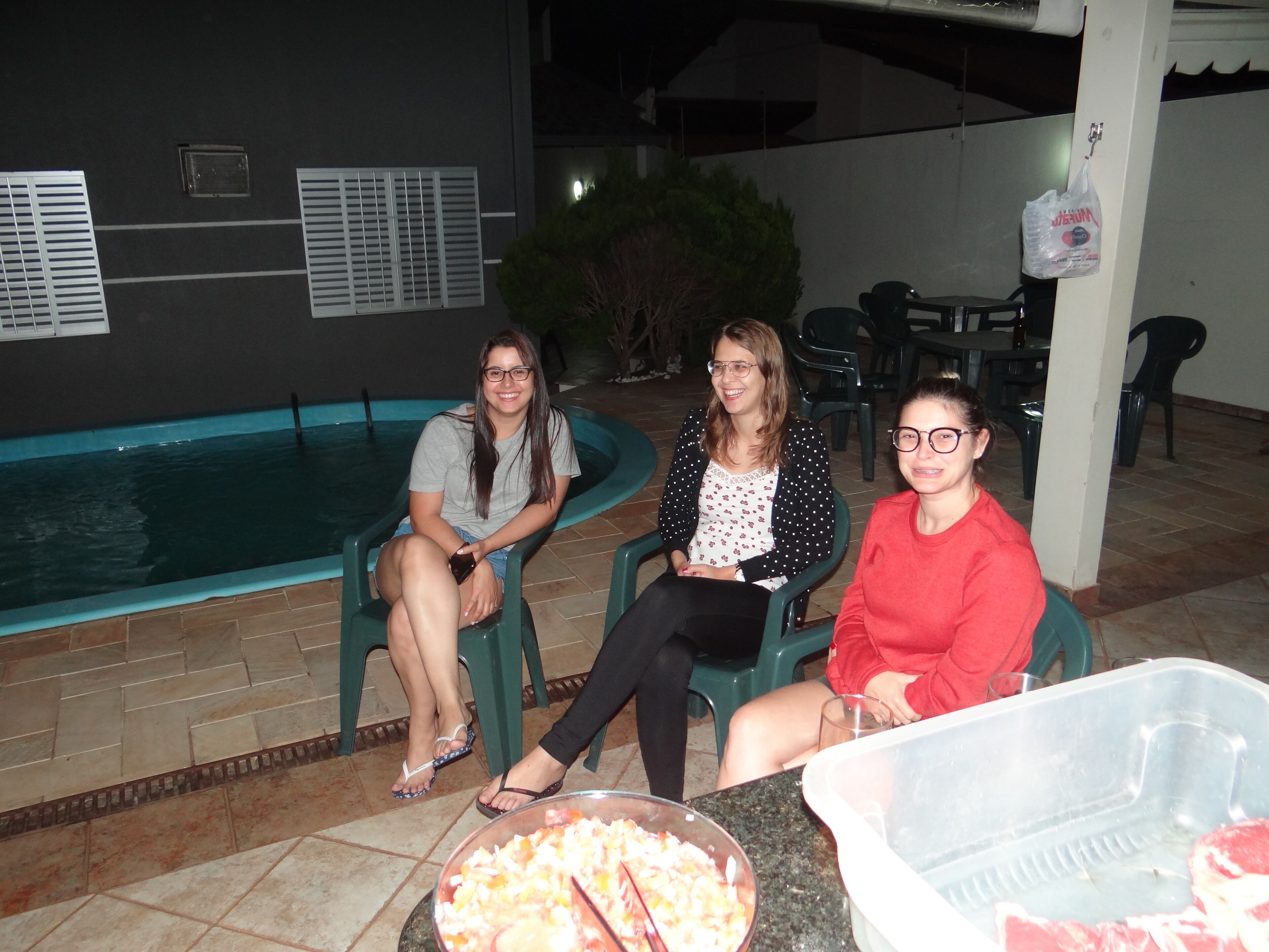 Three people seated by a poolside with a bowl of food and a container of meat on a table.