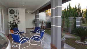 En overdækket terrasse med udendørs møbler, en bil og en have med forskellige planter.