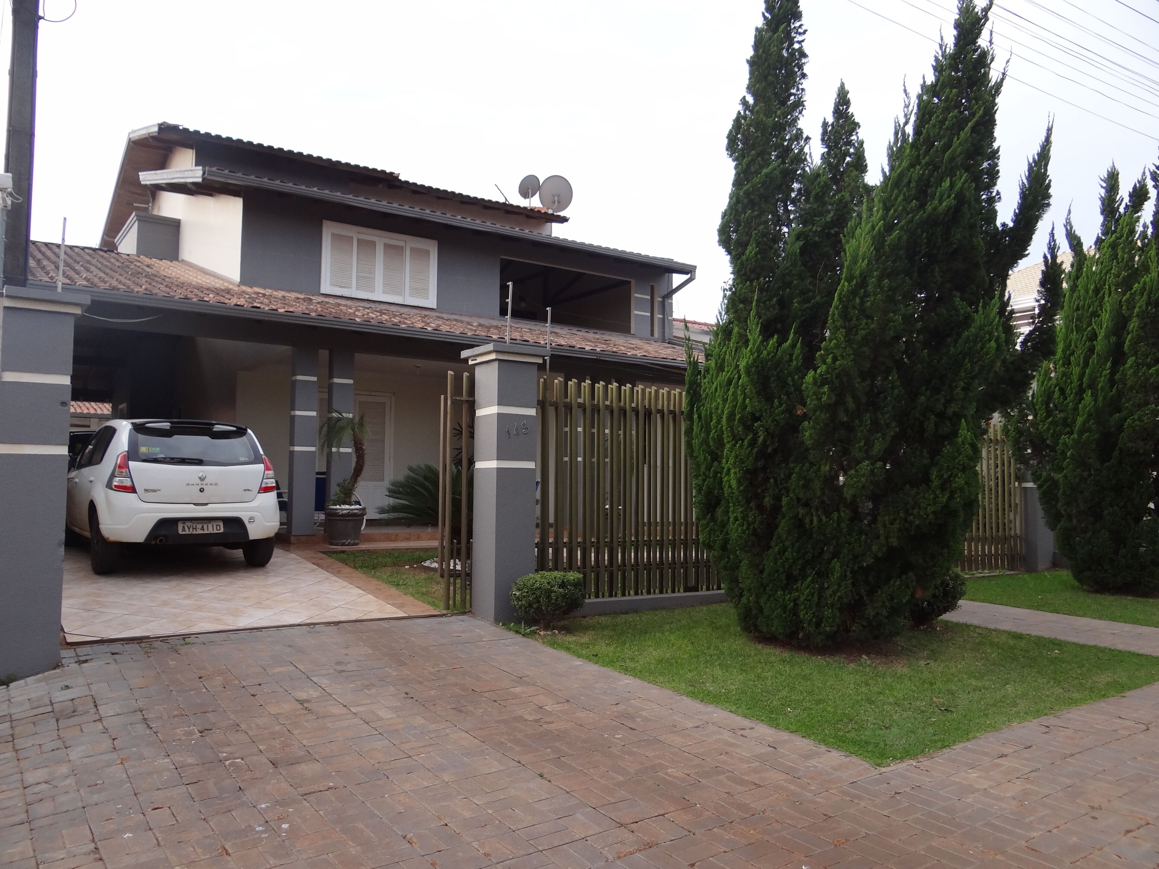 A two-story house with a car parked in the driveway, a fenced yard, and tall evergreen trees.