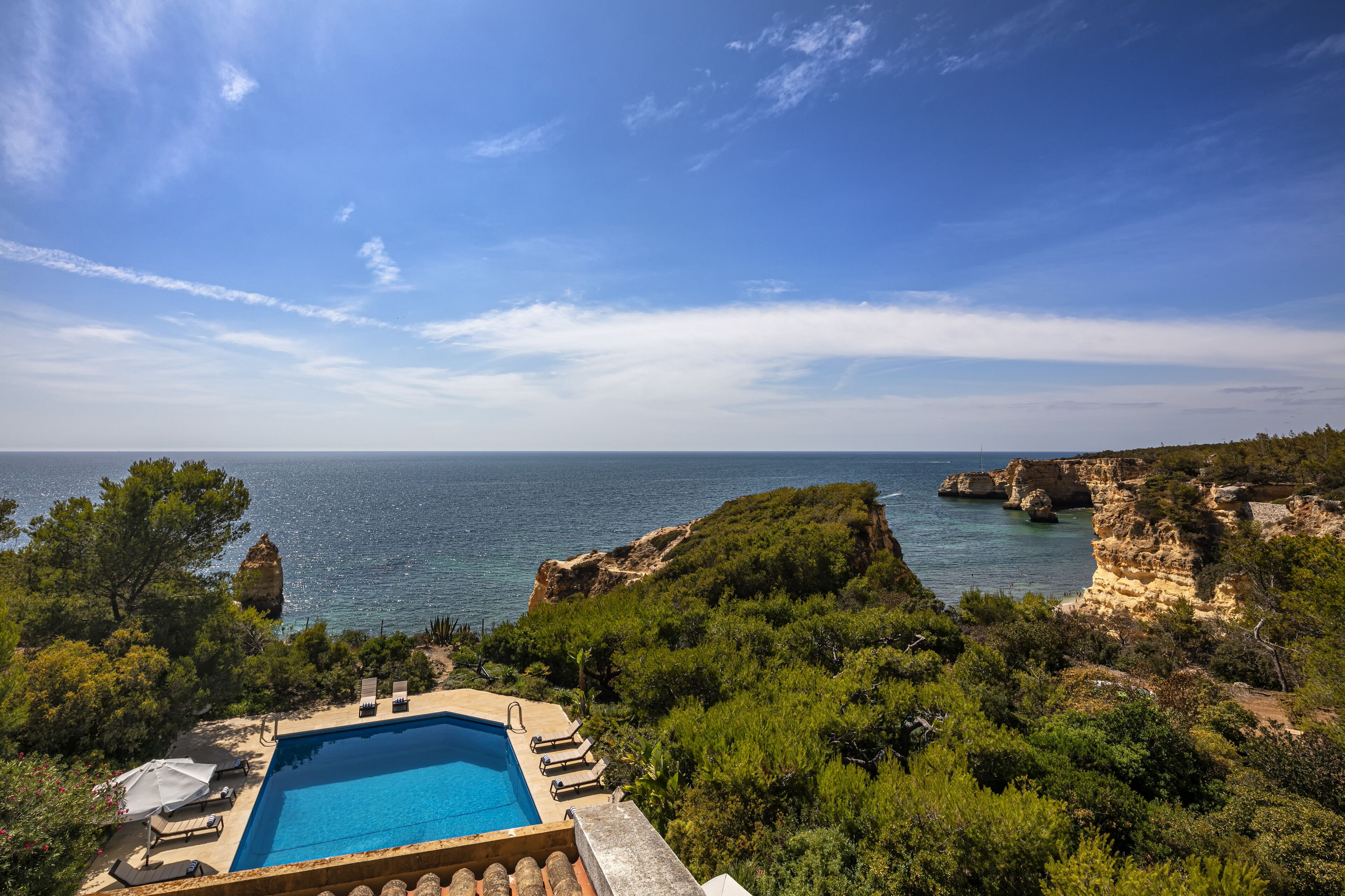 A pool with lounge chairs and umbrellas, surrounded by greenery and overlooking a coastal landscape with cliffs and the sea.