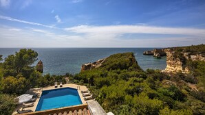 A pool with lounge chairs and umbrellas, surrounded by greenery and overlooking a coastal landscape with cliffs and the sea. - Villa Quinta da Marinha (Carvoeiro)
