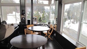 Una habitación acogedora con una mesa redonda y sillas, un sofá, y grandes ventanales con vistas a un paisaje nevado.