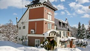 Un edificio de varios pisos con techo inclinado, una entrada destacada con arco decorativo y un paisaje cubierto de nieve.