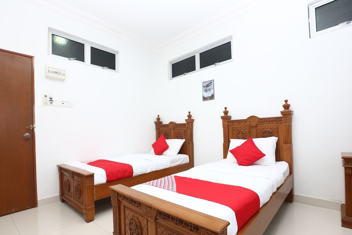 A bedroom with two wooden beds, each with a red and white bedspread and pillows. There are two windows, a door, and a small bedside table.