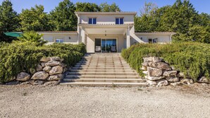 Une maison à deux étages, dotée d’un escalier en pierre menant à l’entrée, entourée de verdure et bordée d’une allée en gravier.