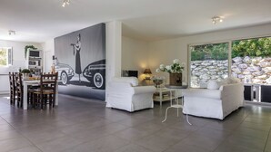Un salon moderne avec un coin repas, agrémenté d’une grande fresque murale, de meubles blancs et d’un mur en pierre à l’extérieur.
