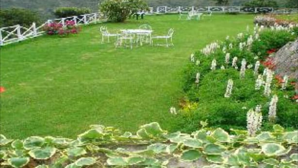 A grassy outdoor area with a white fence, a table and chairs, and a variety of plants. - Mystique Ville (Ootacamund)