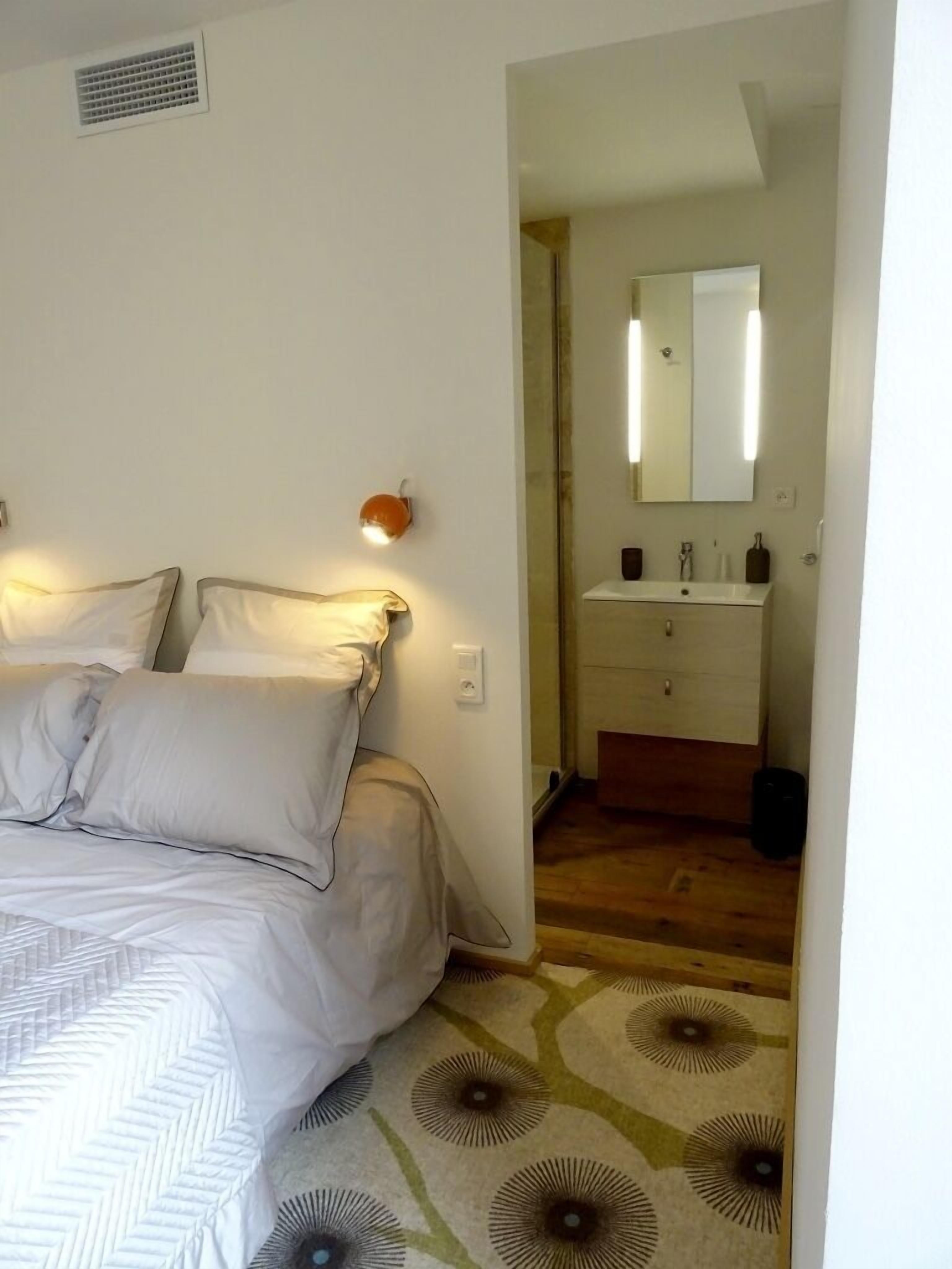 A bedroom with a bed, a nightstand, a wall-mounted lamp, and a bathroom visible through an open door.