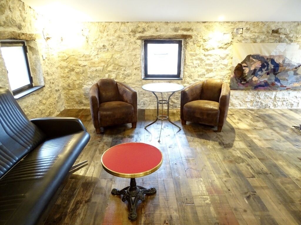 Une pièce avec un mur en pierre, deux fauteuils en cuir, une table ronde rouge et un canapé noir.