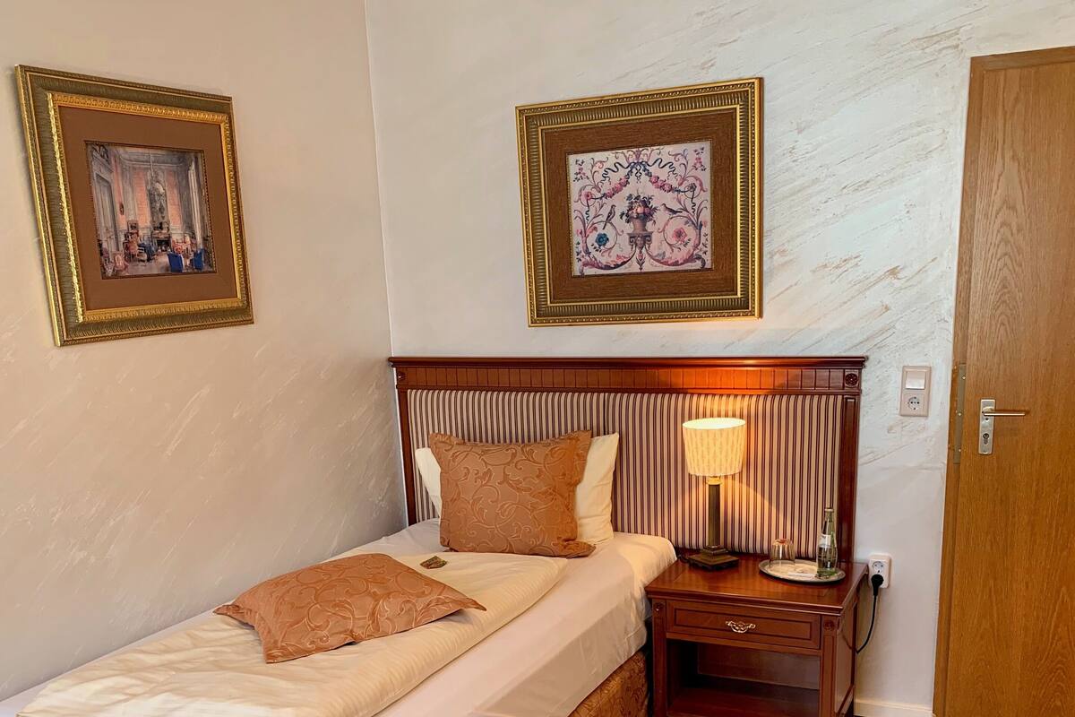 Habitación de hotel ordenada con una cama, una mesita de noche con lámpara, una silla y dos cuadros en la pared.