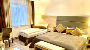 Habitació d'hotel amb llit, taules de nit, una cadira, una taula rodona plena de botelles i una finestra amb cortines.