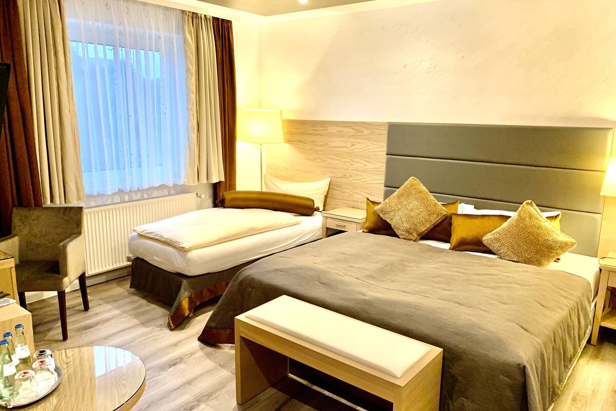 Habitación de hotel con cama, mesitas de noche, una silla, una mesa redonda con botellas y una ventana con cortinas.