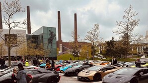Zona aparcament amb diversos cotxes esportius de luxe, un edifici modern amb façana de vidre i algunes persones passant.