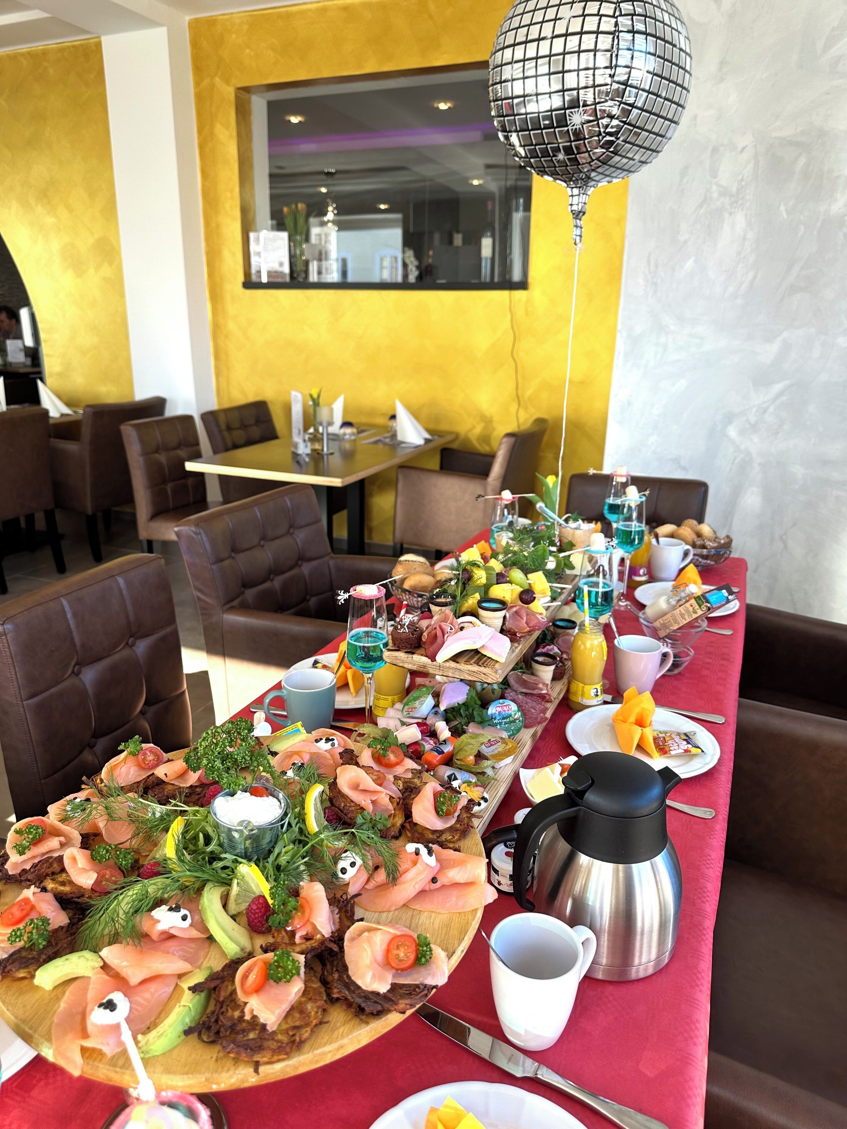 Ein langer Esstisch mit verschiedenen Speisen, darunter Räucherlachs, Käse und Obst, in einem Restaurant mit gelben Wänden und braunen Ledersesseln.