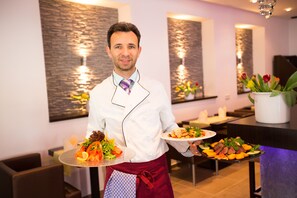 A person in a white chef's coat holding two plates of food in a modern dining area with stone wall decor and a vase of flowers. - Boutique Hotel Goldene Henne (Wolfsburg)