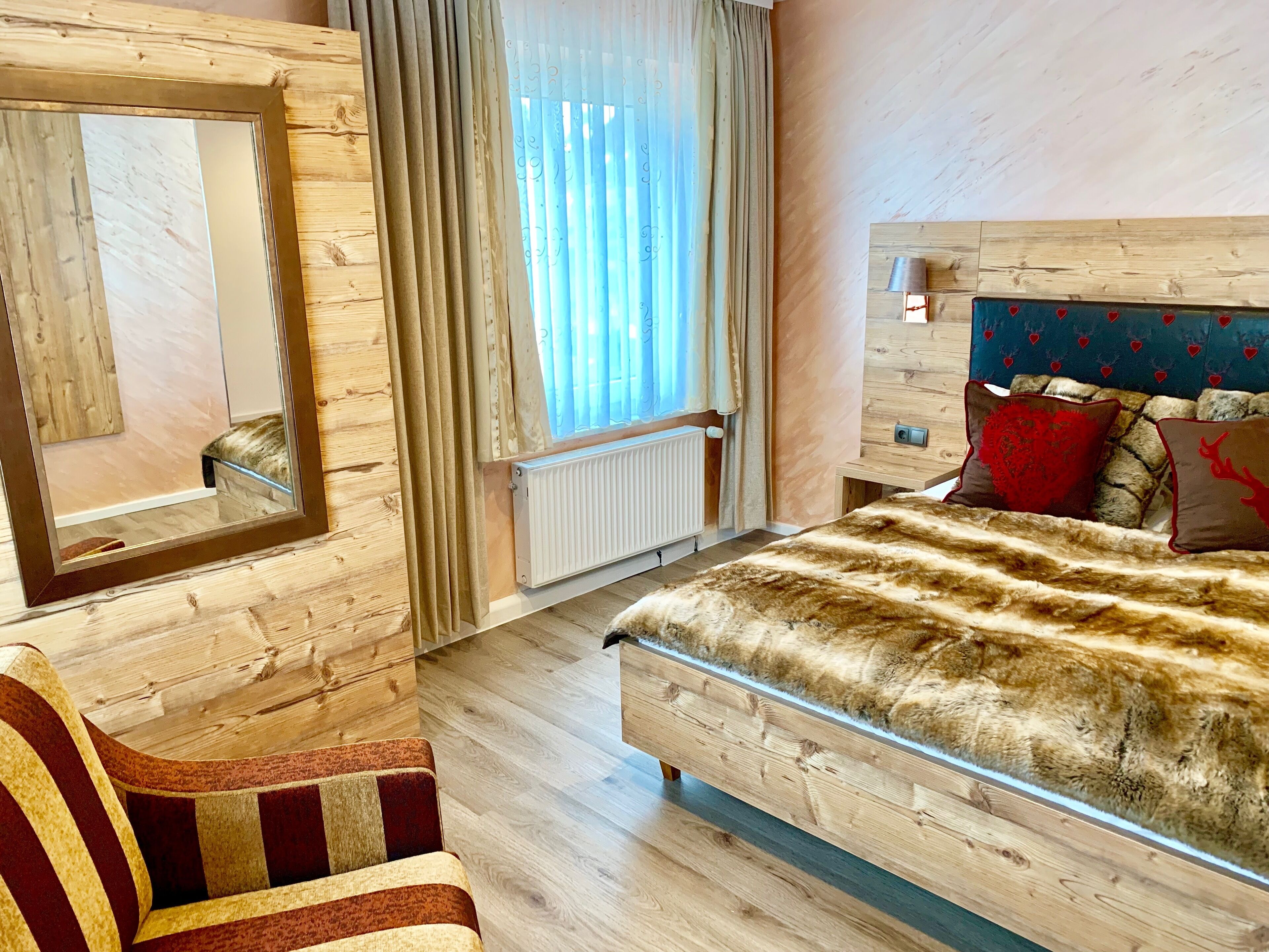 A wooden-paneled room with a bed, a chair, a mirror, and a window with curtains.