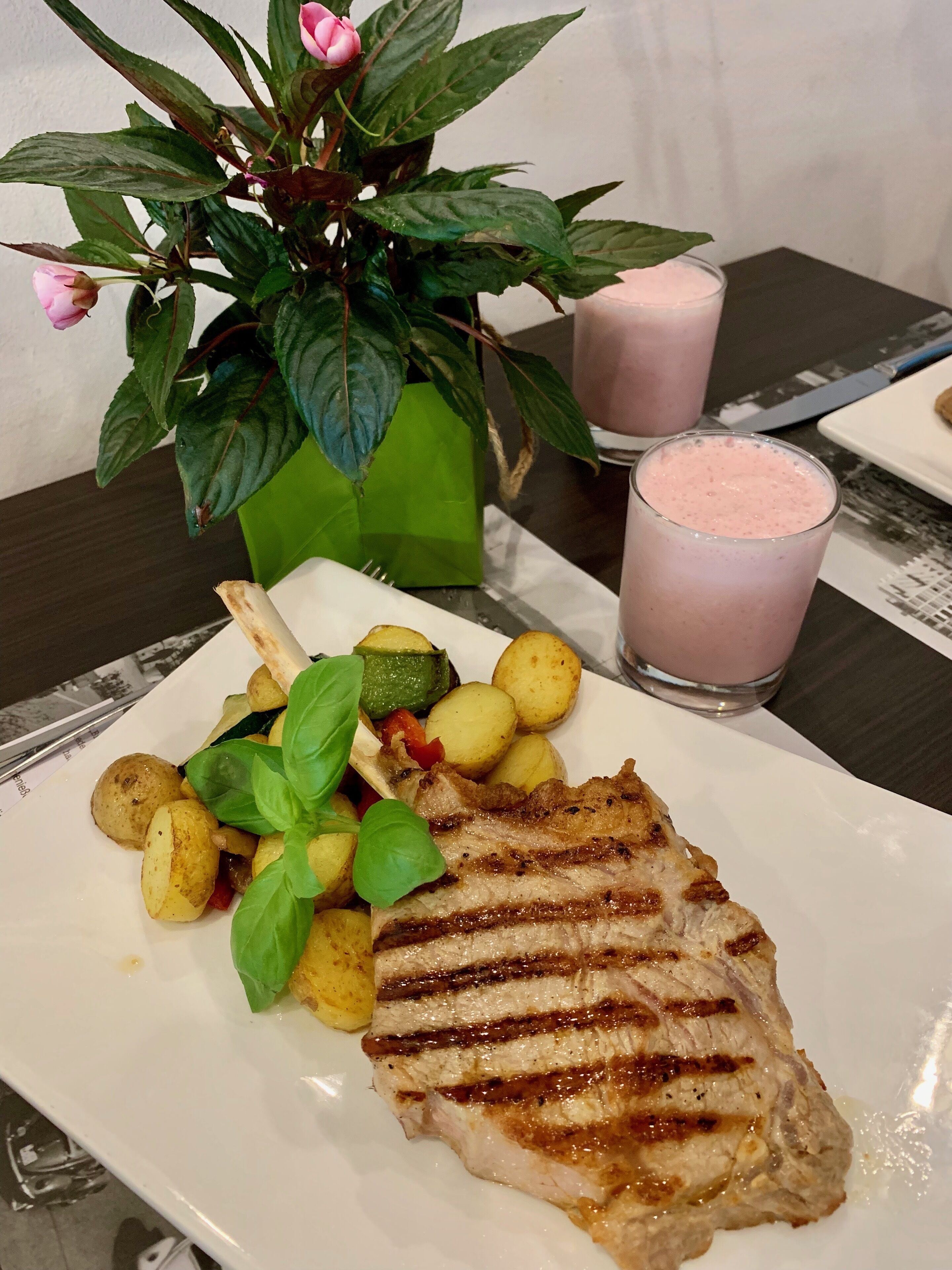 A plate with grilled meat, roasted potatoes, and a green salad, accompanied by a pink drink and a potted plant with a pink flower.