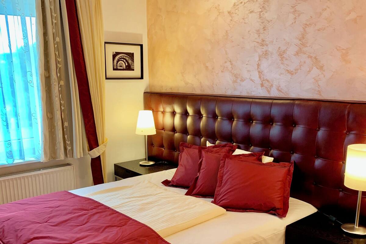 Habitación de hotel con una cama grande, ropa de cama roja y blanca, un cabecero capitonado, una mesita de noche con lámpara y una ventana con cortinas.
