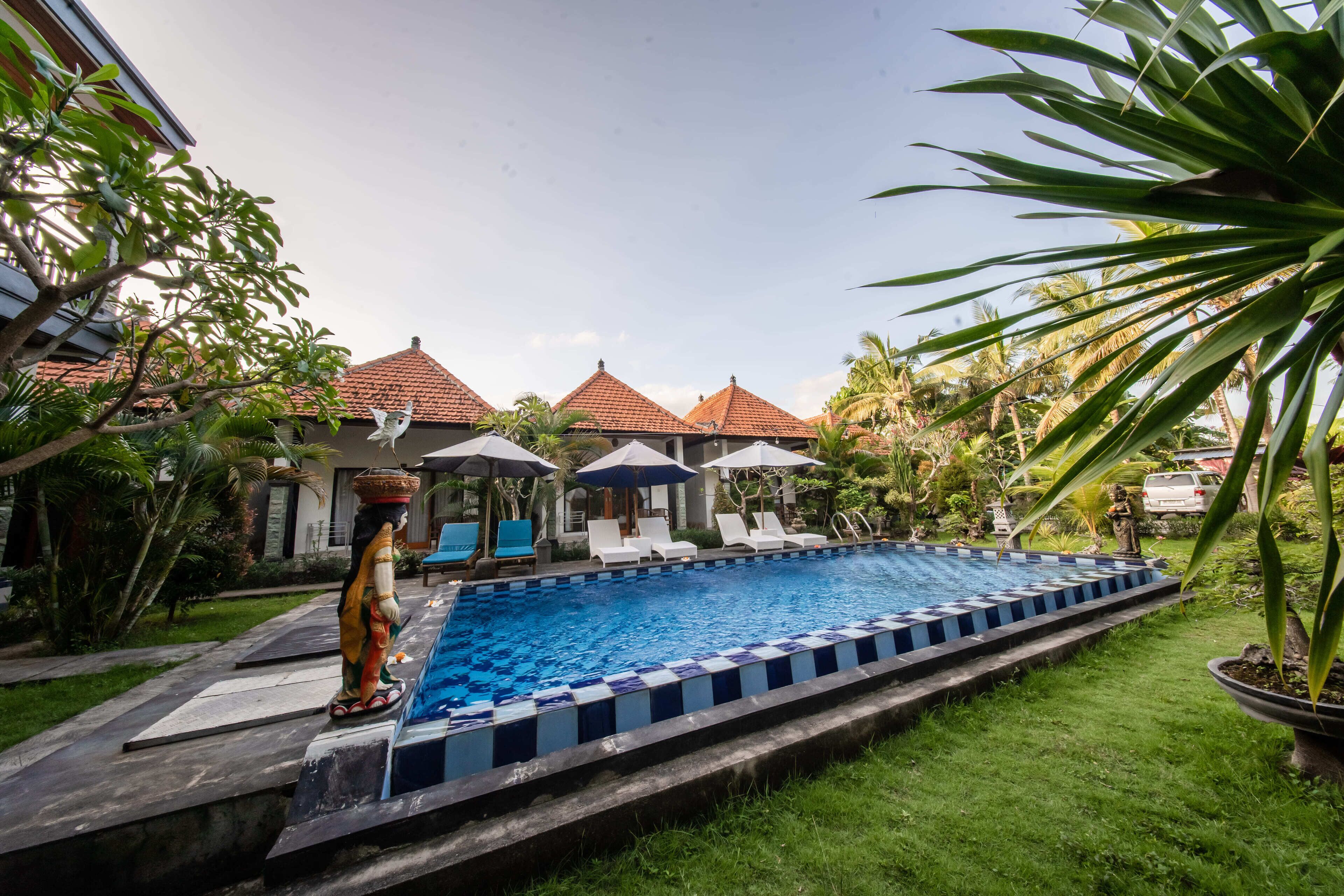 A swimming pool with a tiled deck, surrounded by lounge chairs and umbrellas, with a building with red-tiled roofs in the background.
