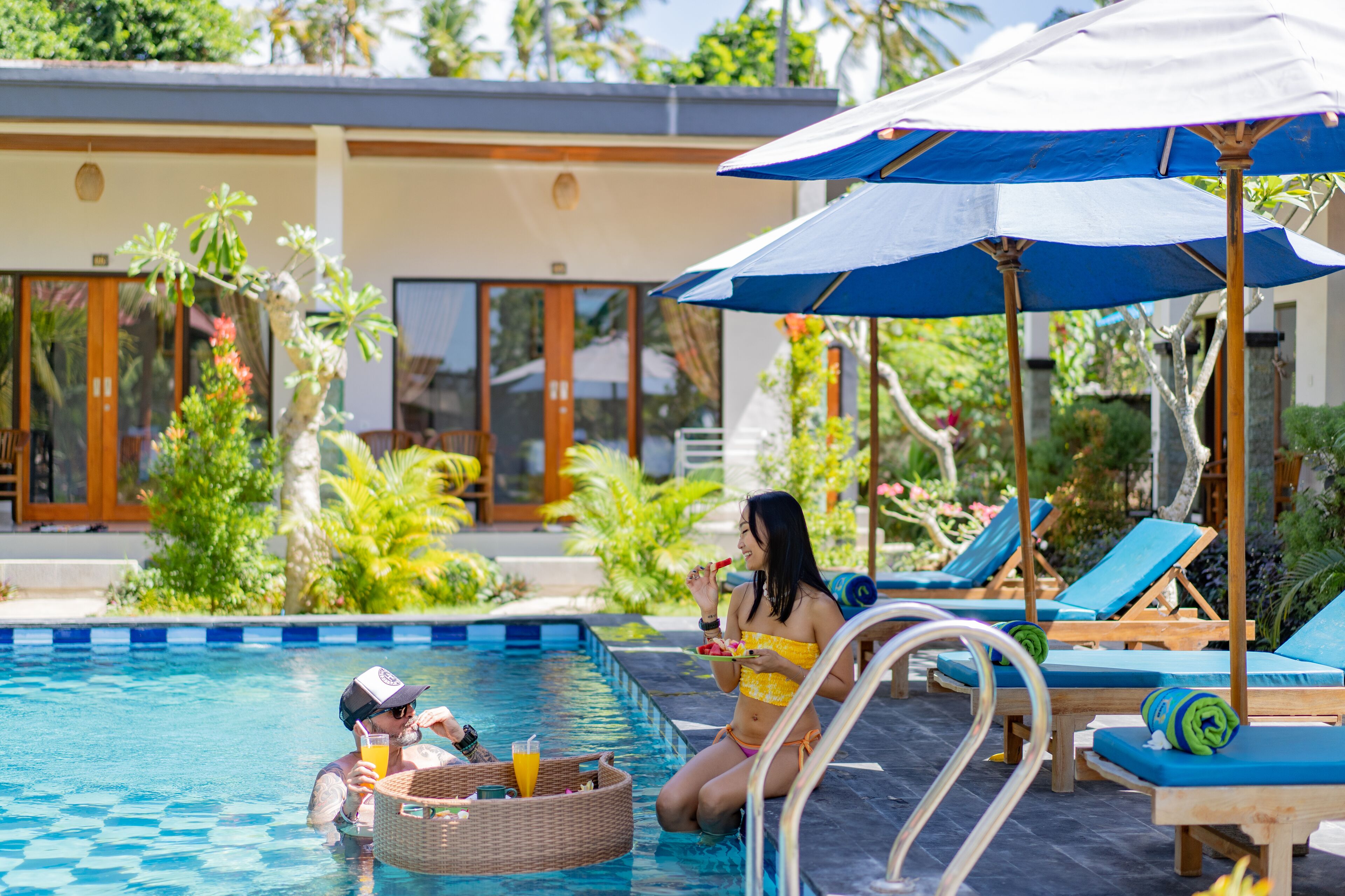 A pool area with lounge chairs and umbrellas, surrounded by greenery and a building with large windows.