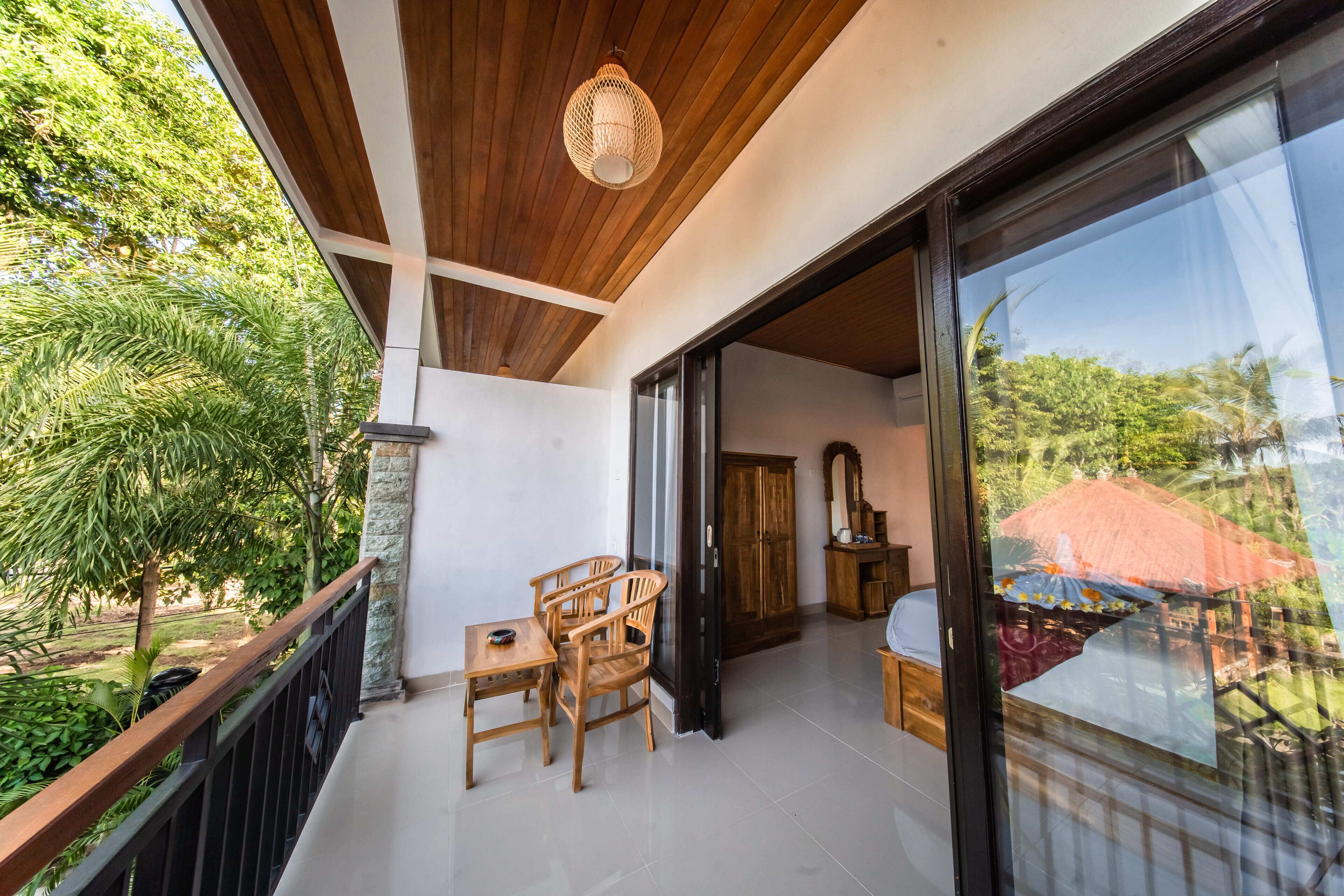 A balcony with a wooden table and chairs, a hanging light fixture, and a view of greenery and a red-roofed building.
